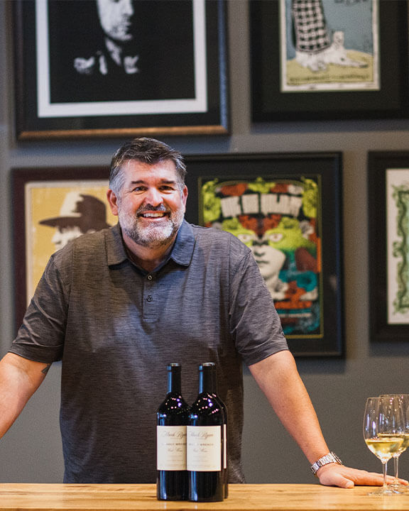 mark mcneilly standing smiling at counter with wine bottles